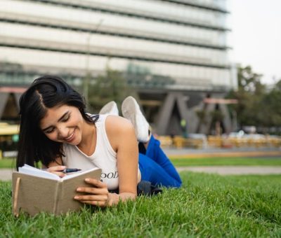 Smiling woman lying on grass reading a notebook in a city park on a sunny day.
