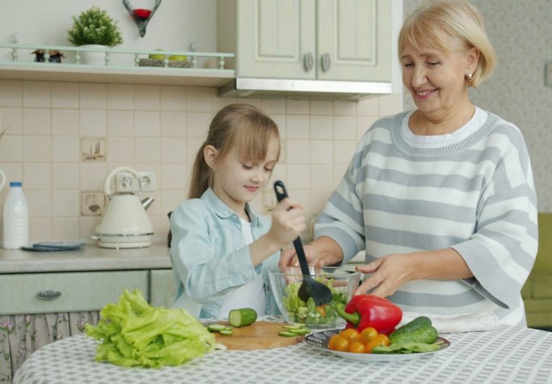 Little girl is cooking salad mixing vegetables in bowl and chatting to cheerful granny in kitchen at home. Modern lifestyle, meals preparation and family concept.
