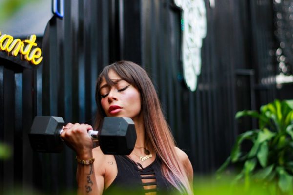 Fit woman with long hair and tattoos lifting a dumbbell with intense focus outdoors.