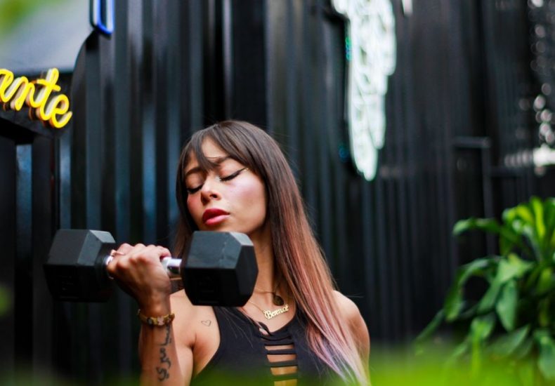 Fit woman with long hair and tattoos lifting a dumbbell with intense focus outdoors.